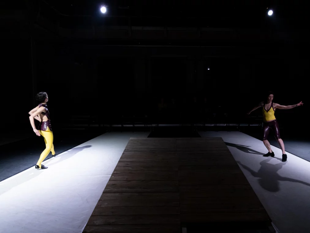 Zwei Personen bewegen sich gemeinsam in einem runden Lichtkegel auf einer Holzfläche. Die Körper sind leicht versetzt zueinander, die Arme heben sich in gespannter, rhythmischer Haltung.