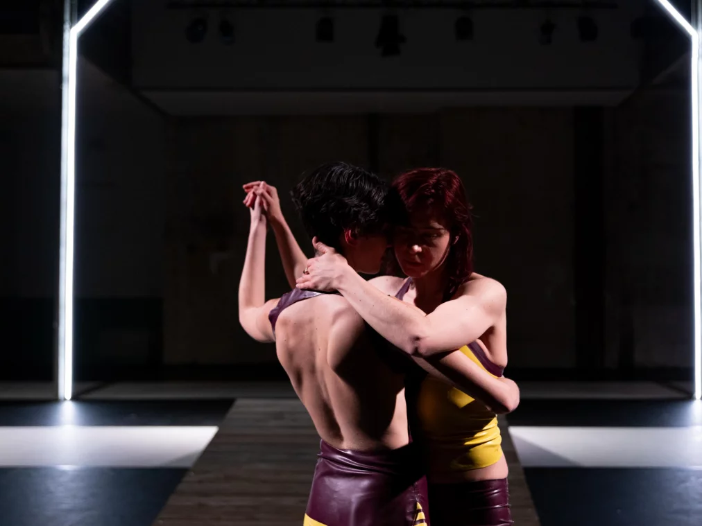 Zwei Personen stehen eng umschlungen im Zentrum eines Bühnenraums. Die Oberkörper berühren sich, die Arme sind ineinander verschränkt. Der Raum ist symmetrisch aufgebaut, mit einer architektonischen Lichtstruktur im Hintergrund, die die intime Nähe der Szene betont.