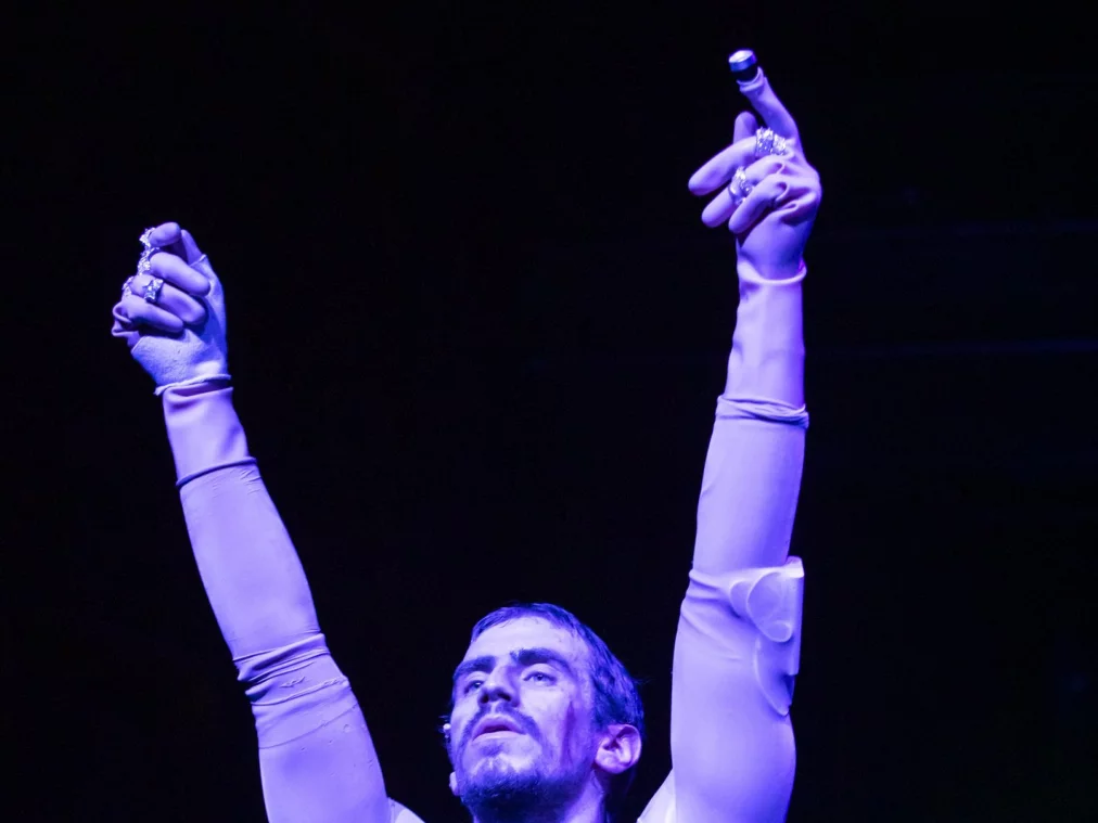 Nahaufnahme einer Person auf einer Bühne, die beide Arme gestreckt nach oben hebt. Die Person trägt ein eng anliegendes, helles Ganzkörperkostüm mit Handschuhen und Schmuck an den Fingern. Violettes Bühnenlicht beleuchtet Gesicht und Oberkörper, während der Hintergrund dunkel bleibt. Ein kleines Funkgerät ist am Kostüm befestigt.