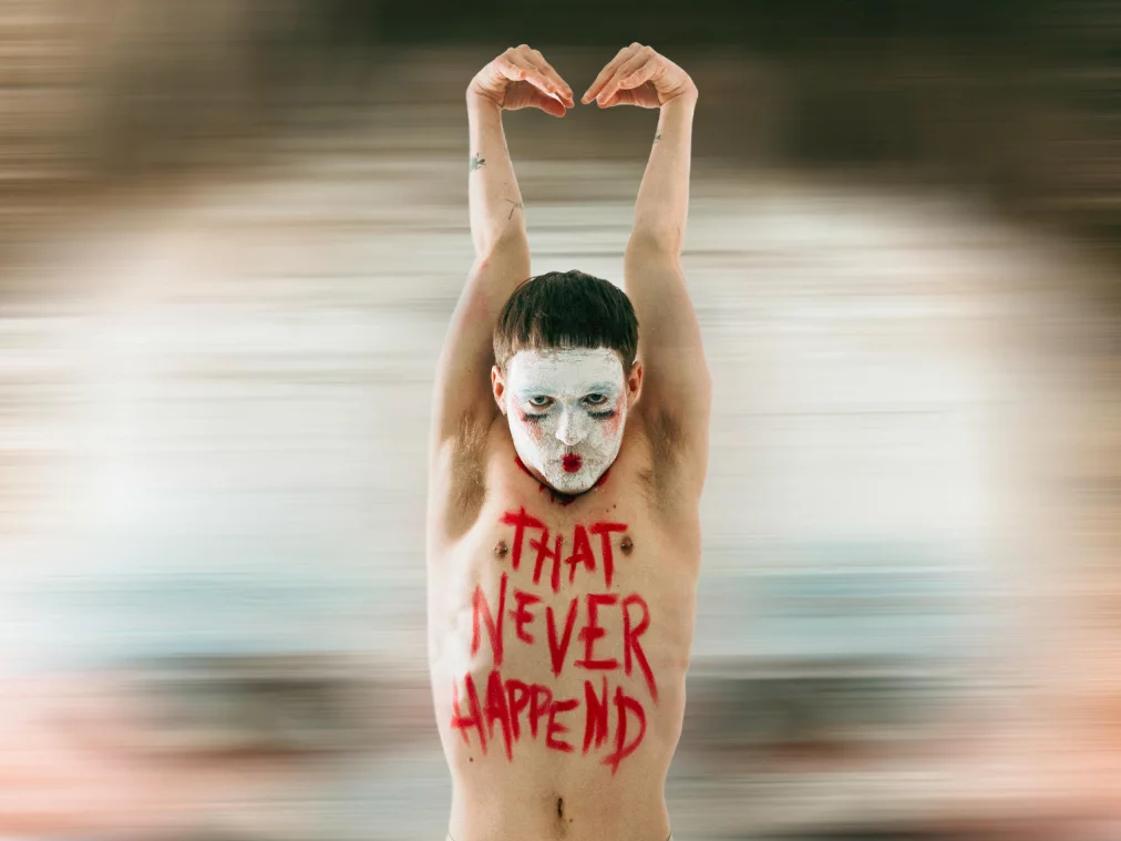 Eine Person steht frontal vor einem verschwommenen, rot-beigen Hintergrund. Das Gesicht ist weiß geschminkt mit stark betonten Augen und roten Lippen. Auf dem Oberkörper steht in roter Farbe „THAT NEVER HAPPEND“. Die Arme sind über dem Kopf nach oben gestreckt, die Hände berühren sich. Die Person trägt enganliegende graue Leggings.