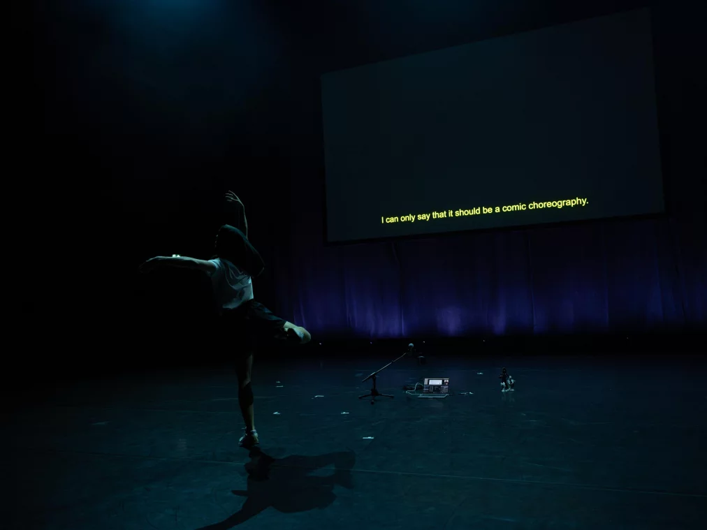 Eine Person bewegt sich auf einer dunklen Bühne im seitlichen Licht. Im Hintergrund hängt eine große Leinwand mit gelber Schrift. Auf dem Boden stehen ein Laptop, ein Mikrofonständer und kleine Figure