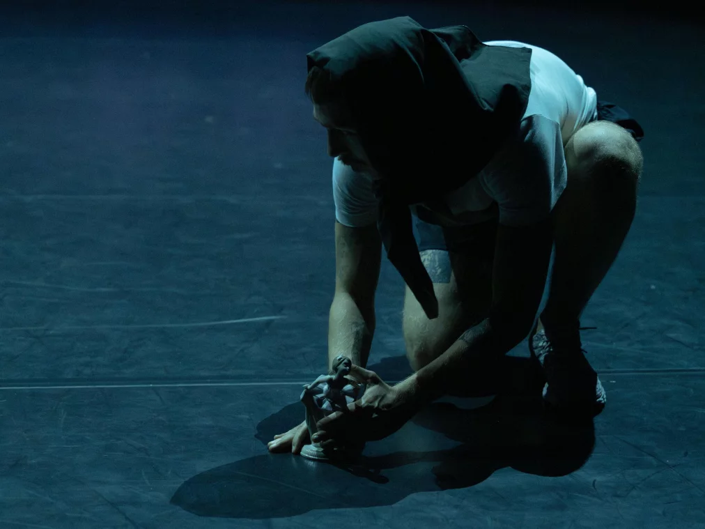 Eine Person hockt auf einer dunklen Bühne und hält eine kleine Figur in den Händen. Die Person trägt ein helles T-Shirt, Shorts und ein dunkles Tuch über dem Kopf. Kühles, gerichtetes Licht erzeugt starke Schatten auf dem Boden.