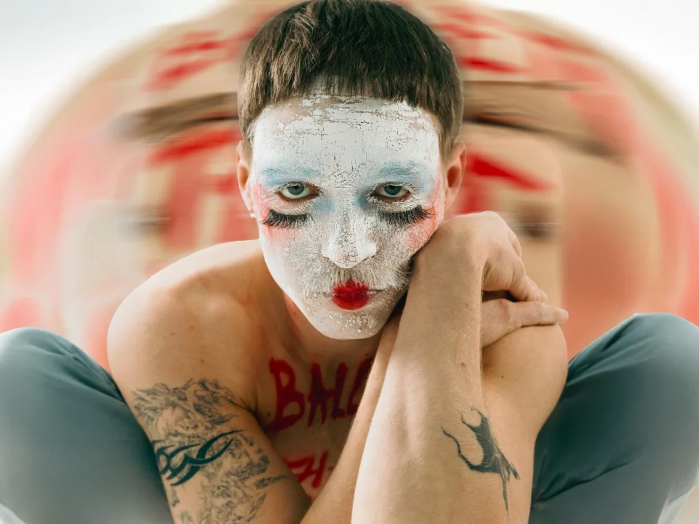 Porträt einer Person mit kurz geschnittenen Haaren und weiß geschminktem Gesicht. Die Person sitzt mit angewinkelten Armen und stützt den Kopf auf die Hände. Rote Lippen, betonte Wimpern und sichtbare Tattoos an den Armen prägen das Erscheinungsbild. Im Hintergrund befindet sich eine unscharfe, rot-beige Farbfläche.