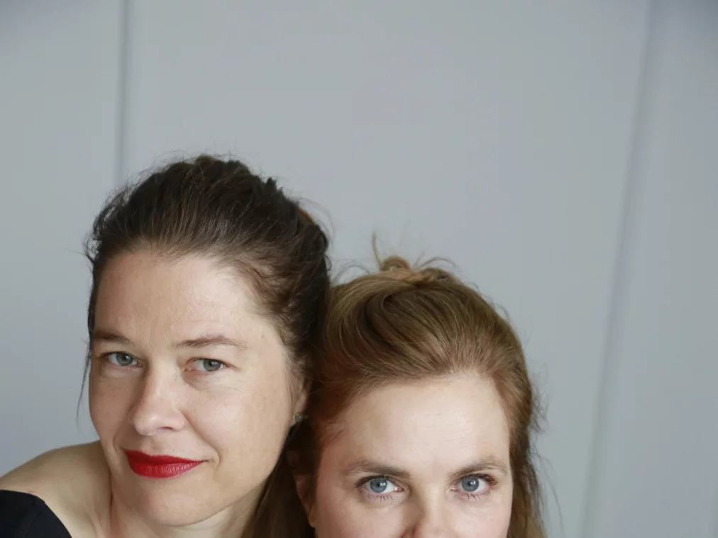 Zwei Personen sitzen eng nebeneinander vor einem hellen Hintergrund und schauen in die Kamera. Beide tragen schwarze Oberteile.