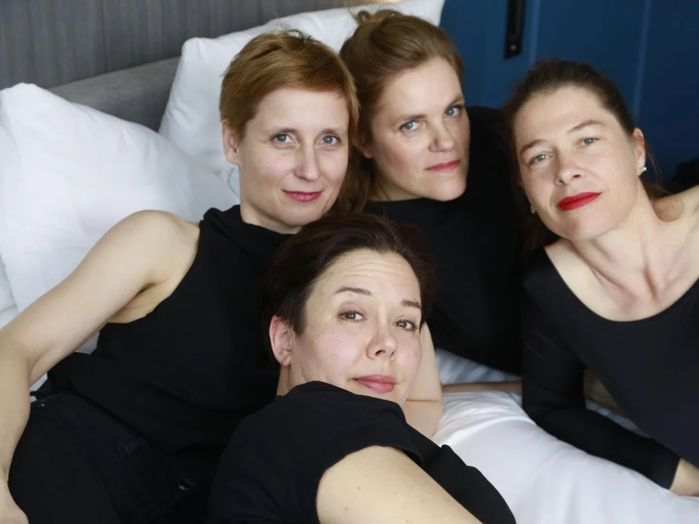 Vier Personen liegen eng nebeneinander auf einem Bett und blicken in die Kamera. Alle tragen schwarze Oberteile.