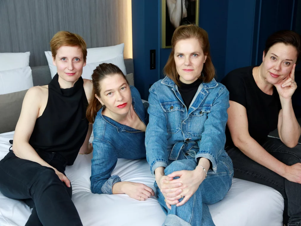 Vier Personen sitzen nebeneinander auf einem Bett in einem Hotelzimmer. Sie schauen in die Kamera und tragen schwarze Kleidung und eine Jeansjacke.