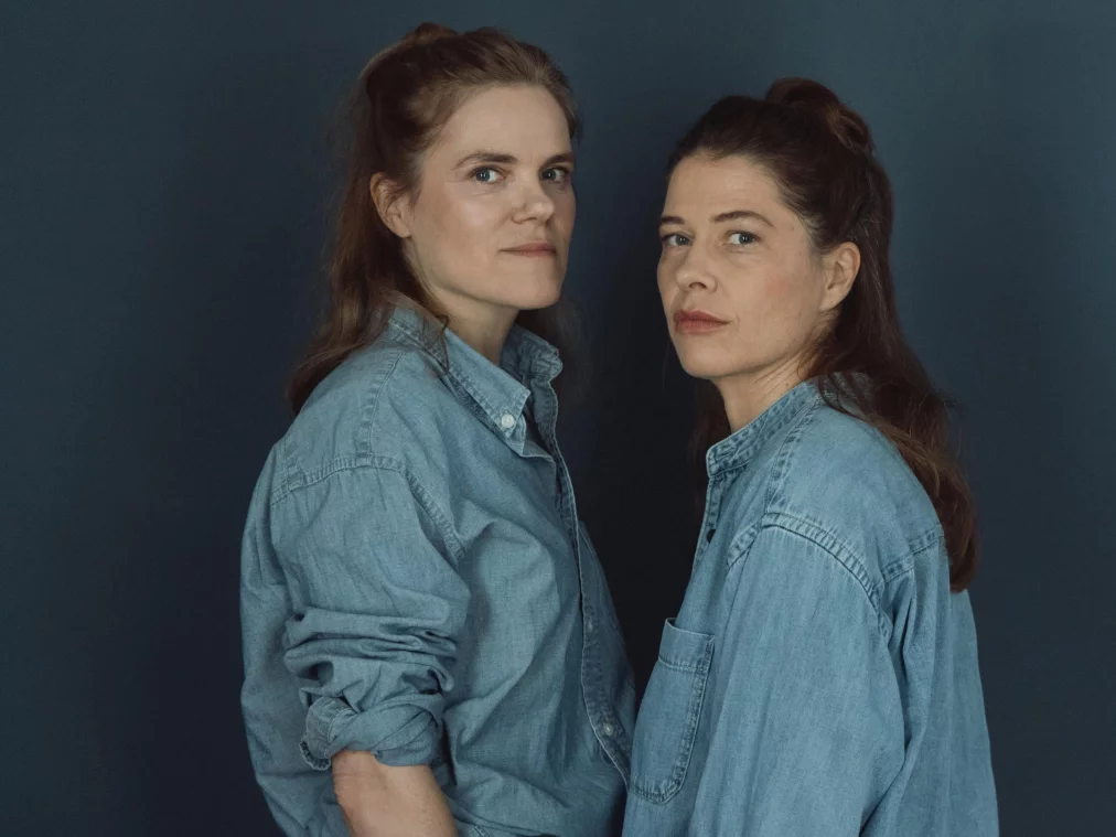 Zwei Personen stehen seitlich zueinander vor einem dunklen Hintergrund und schauen in die Kamera. Beide tragen Jeanshemden.