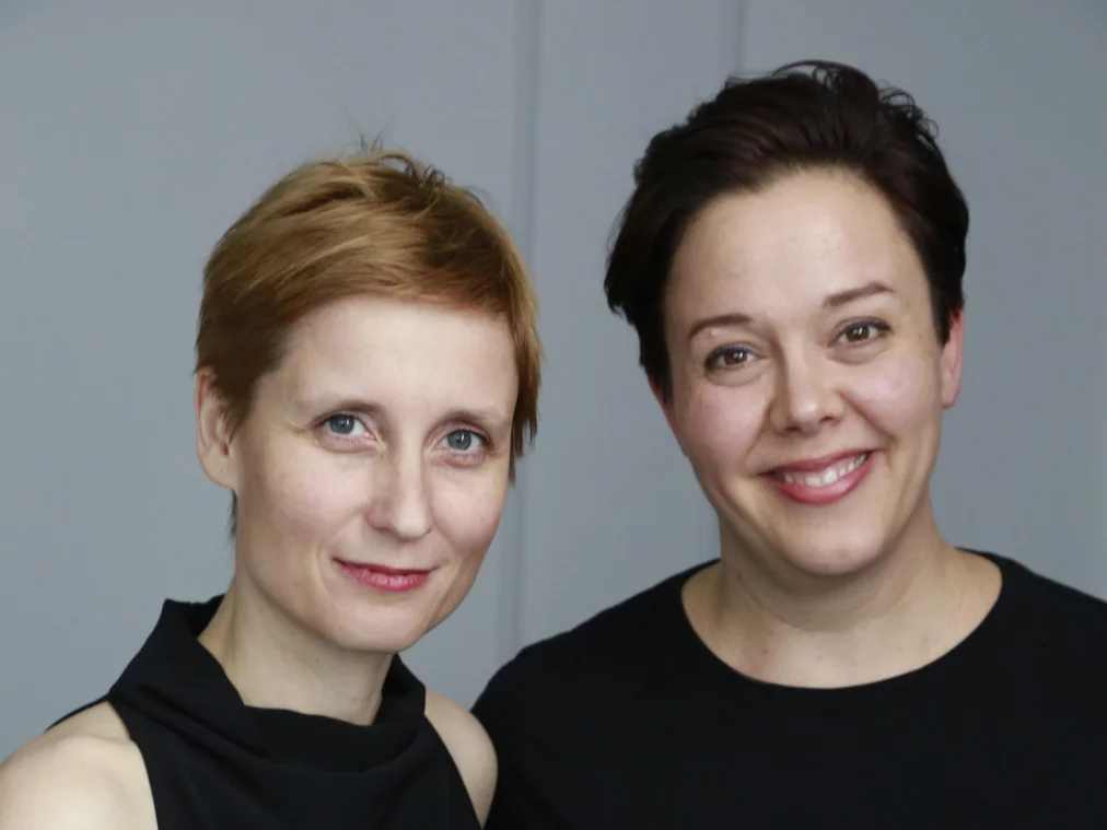 Zwei Menschen stehen nebeneinander vor hellem Hintergrund und blicken in die Kamera. Beide tragen schwarze Oberteile. Eine Person hat kurzes blondes Haar, die andere Person kurzes dunkles Haar.