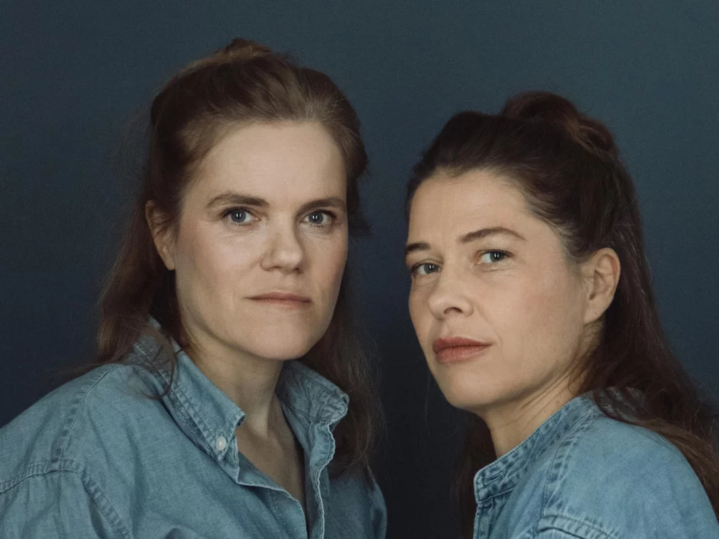 Zwei Personen stehen seitlich zueinander vor einem dunklen Hintergrund und schauen in die Kamera. Beide tragen Jeanshemden.