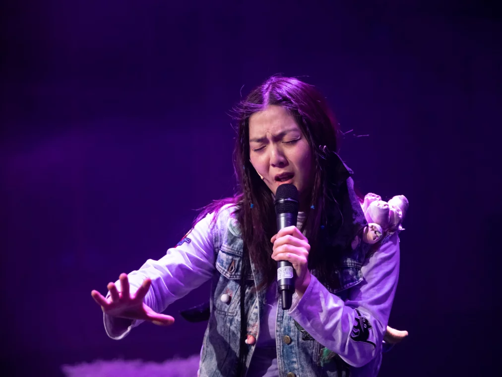 Person singt mit geschlossenen Augen in ein Handmikrofon, violettes Bühnenlicht beleuchtet Gesicht und Oberkörper.