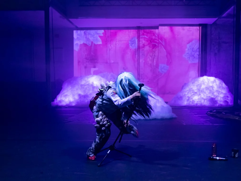 Person mit langer, hellblauer Perücke beugt sich singend über ein Mikrofon auf einer Bühne mit pink-violetter Beleuchtung und wolkenartigen Bühnenobjekten im Hintergrund.