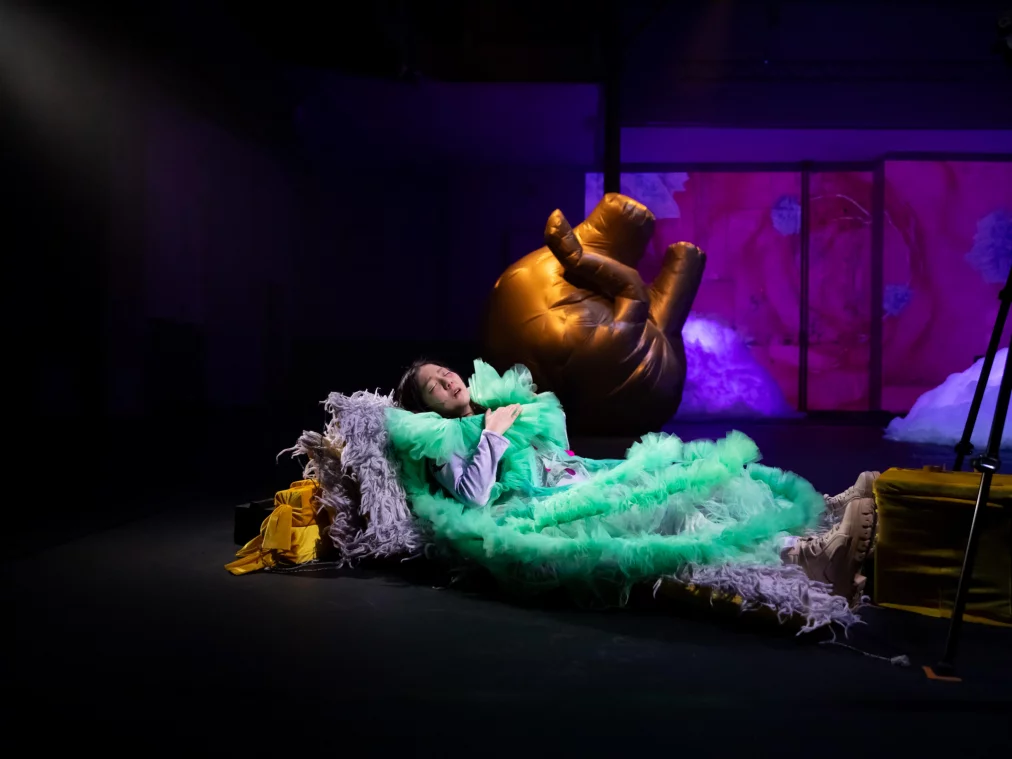 Person in grünem Tüllkostüm liegt auf der Bühne, umgeben von Requisiten, während im Hintergrund ein großes aufblasbares Objekt und farbig beleuchtete Bühnenbilder zu sehen sind.