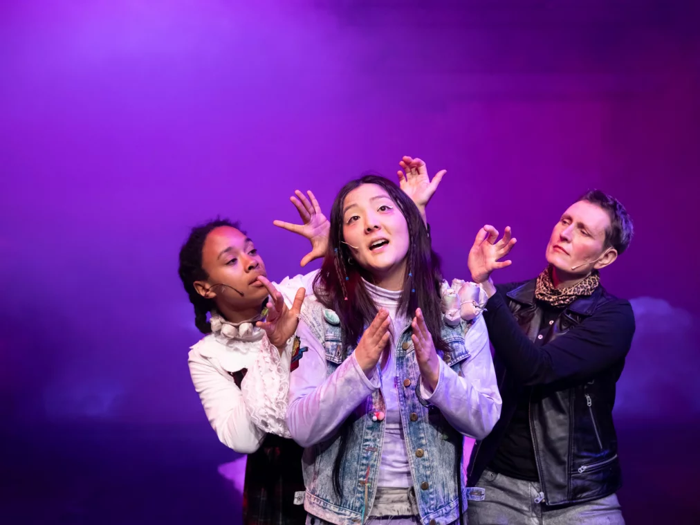 Drei Personen stehen auf einer Bühne, die mittlere Person singt, während zwei weitere Personen gestisch hinter ihr agieren, violette Beleuchtung im Hintergrund.