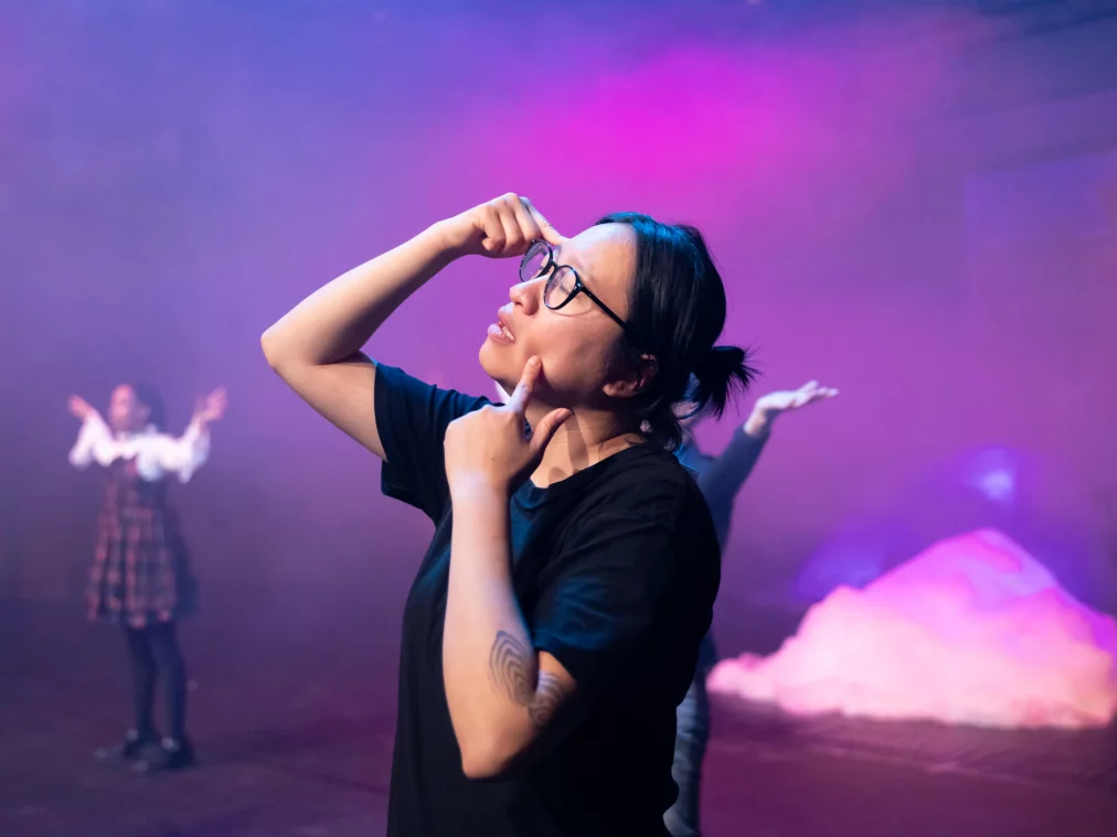 Person mit Brille zeigt mit den Fingern auf Gesicht und Kopf, während zwei weitere Personen unscharf im Hintergrund auf einer violett beleuchteten Bühne stehen.