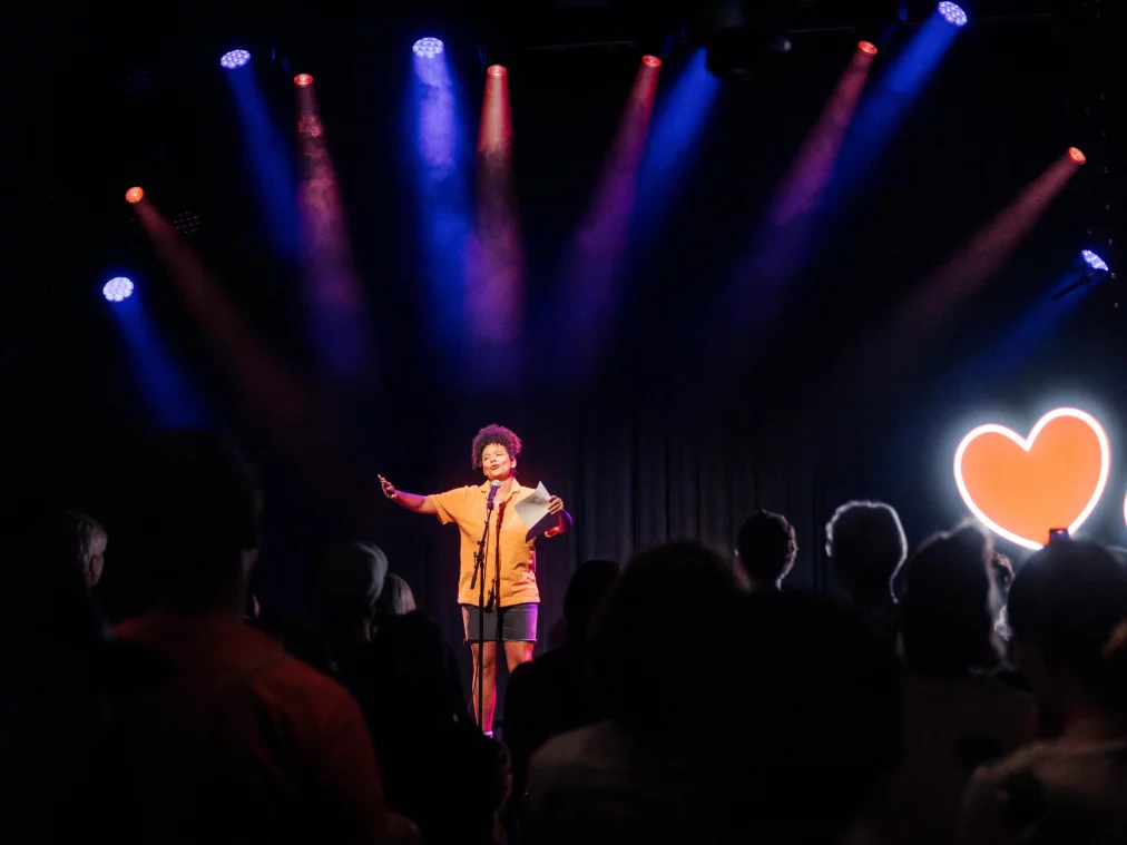 Eine Person steht auf einer beleuchteten Bühne vor einem Publikum und hält ein Blatt Papier in einer Hand, während die andere offen in den Raum gestreckt ist. Über der Bühne hängen farbige Scheinwerfer, seitlich leuchten zwei herzförmige Lichtobjekte.