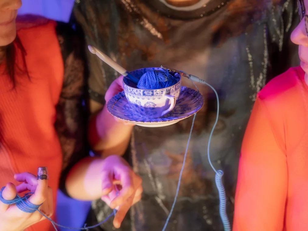 Mehrere Personen stehen eng beieinander. Eine Person hält eine blau-weiß gemusterte Teetasse mit einer Kugel blauer Wolle darin, auf einer Untertasse. Blaue Fäden verbinden die Hände der Personen miteinander.