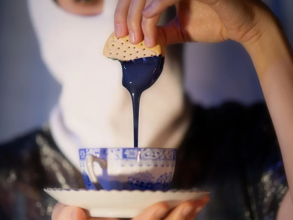 Eine Person hält eine blau-weiß gemusterte Teetasse auf einer Untertasse. Ein Keks wird in eine dickflüssige, dunkelblaue Substanz getaucht, die langsam in die Tasse zurückläuft. Der Oberkörper der Person ist unscharf im Hintergrund zu sehen.