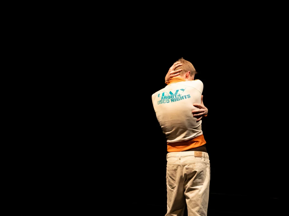 Eine einzelne Person steht vor schwarzem Hintergrund und umschließt den eigenen Oberkörper mit den Armen, eine Hand liegt am Hinterkopf. Das helle Oberteil mit orangefarbenen Akzenten hebt sich stark vom dunklen Raum ab.