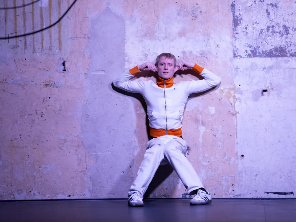 Eine Person sitzt mit dem Rücken an einer rauen, hellen Wand und beugt die Arme angewinkelt nach oben, die Hände nahe den Ohren. Die Person trägt einen hellen Trainingsanzug mit orangefarbenen Details und blickt frontal in die Kamera.