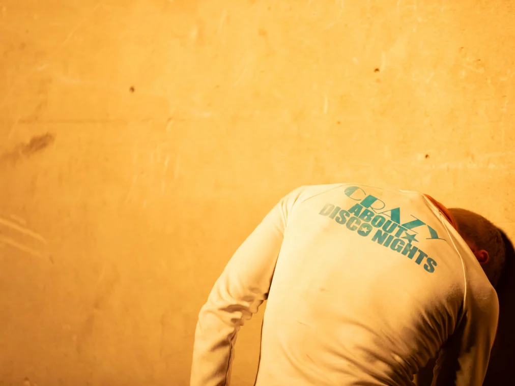 Eine Person steht dicht vor einer gelblich beleuchteten Wand und beugt den Oberkörper nach vorn, sodass der Kopf nicht sichtbar ist. Auf dem Rücken des hellen Oberteils ist der Schriftzug „CRAZY ABOUT DISCO NIGHTS“ zu lesen.
