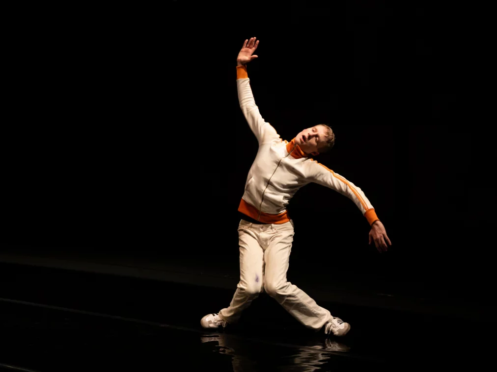 Eine Person bewegt sich seitlich über eine dunkle, glänzende Bühne, ein Arm ist weit nach oben gestreckt, der andere hängt locker nach unten. Das Licht betont die schräge Körperhaltung und erzeugt Spiegelungen auf dem Boden.