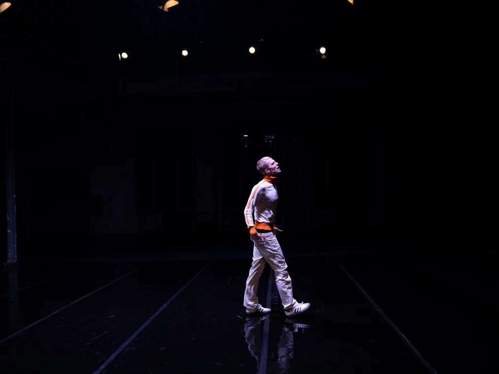 Eine Person steht allein auf einer großen dunklen Bühne und blickt nach oben zu mehreren kleinen Deckenleuchten. Der Körper ist seitlich ausgerichtet, das Licht zeichnet klare Konturen vor dem schwarzen Hintergrund.