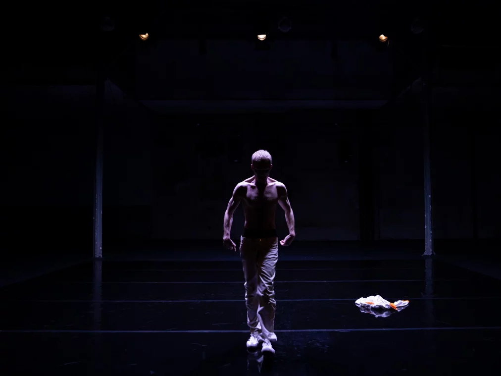 Eine Person mit freiem Oberkörper geht langsam über eine dunkle Bühne auf die Kamera zu. Von oben fällt ein konzentrierter Lichtkegel auf den Körper, während der Raum ringsum im Schatten bleibt. Rechts liegt ein helles Kleidungsstück auf dem Boden.