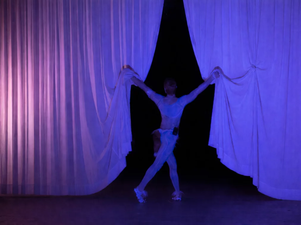 Eine Person steht mittig auf einer Bühne zwischen zwei großen, halb geöffneten, weißen Vorhängen. Die Person hält mit beiden Armen jeweils eine Seite des Stoffes auseinander. Sie trägt ein enges, helles Bühnenkostüm und Turnschuhe. Das Licht ist blau-violett gefärbt und lässt den Hintergrund hinter den Vorhängen dunkel erscheinen.
