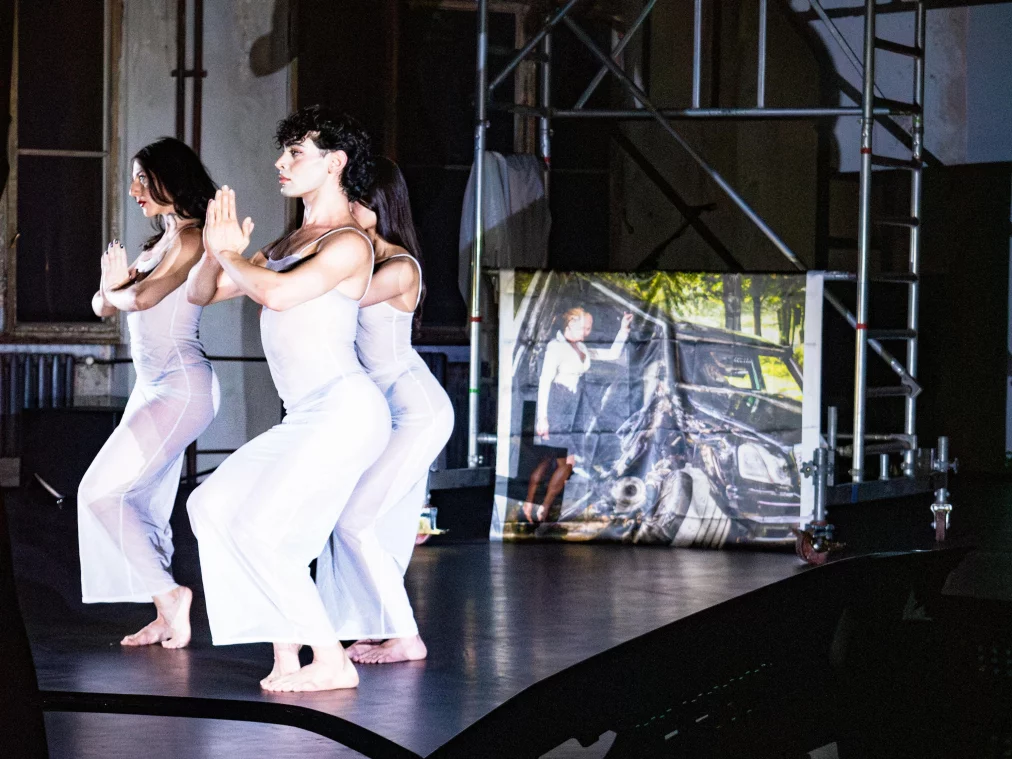 Drei Personen in transparenten weißen Kostümen bewegen sich synchron auf einer erhöhten Bühnenfläche vor einer Gerüststruktur und einer Bildprojektion mit einem Autounfallmotiv.