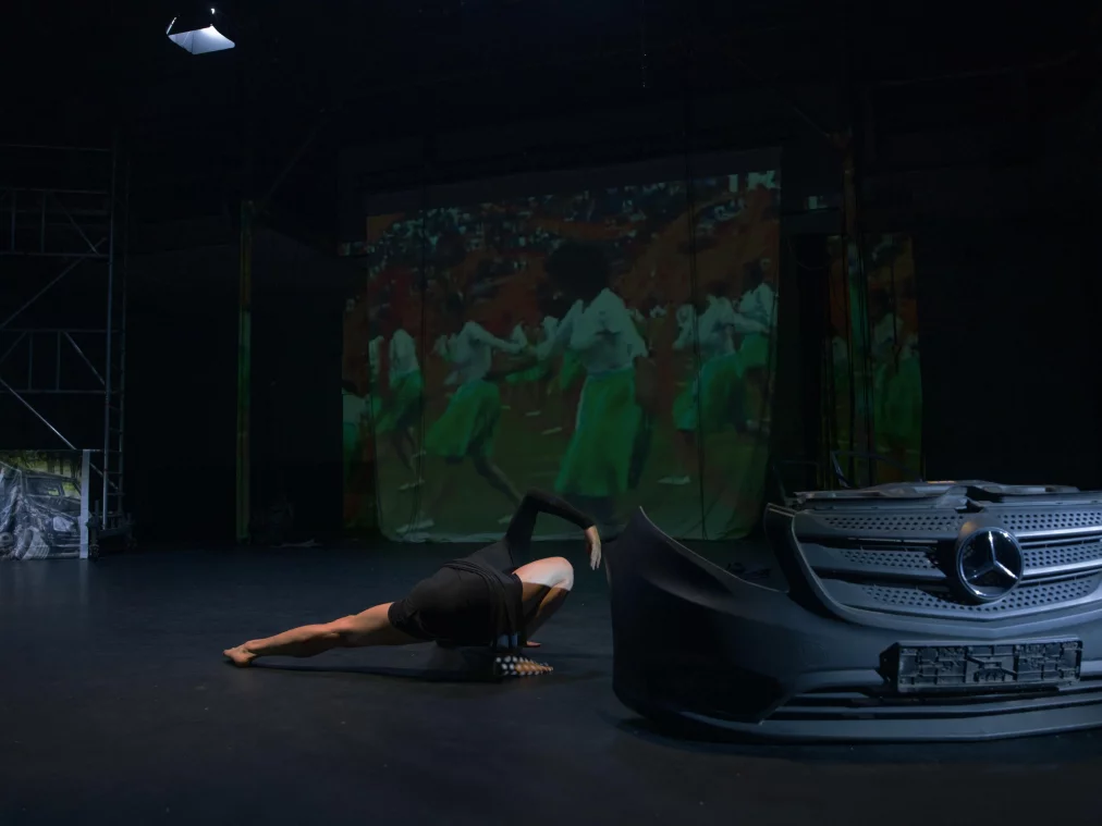 Eine Person in schwarzem Kostüm liegt in einer tiefen, seitlich gestreckten Bodenpose neben einer abmontierten Auto-Front auf einer Bühne; im Hintergrund ist eine Videoprojektion mit tanzenden Menschen zu sehen.