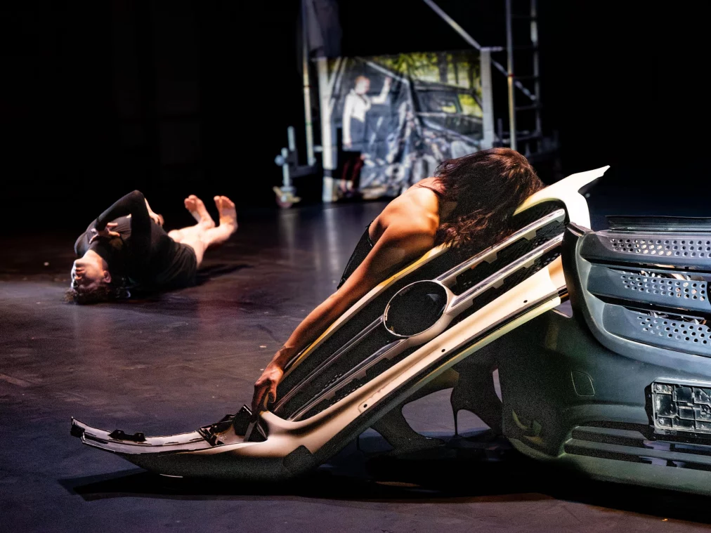 Zwei Personen liegen erschöpft auf dem Bühnenboden neben und auf abgenommenen Autoteilen; im Hintergrund sind Bühnenelemente und Projektionen sichtbar.