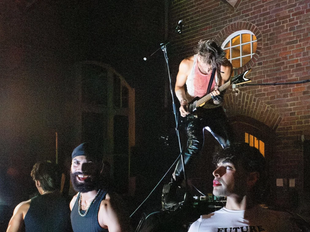 Mehrere Personen posieren nachts um ein Auto in einem Innenhof; eine Person steht auf dem Fahrzeug und spielt Gitarre, während andere Personen um das Auto verteilt stehen.