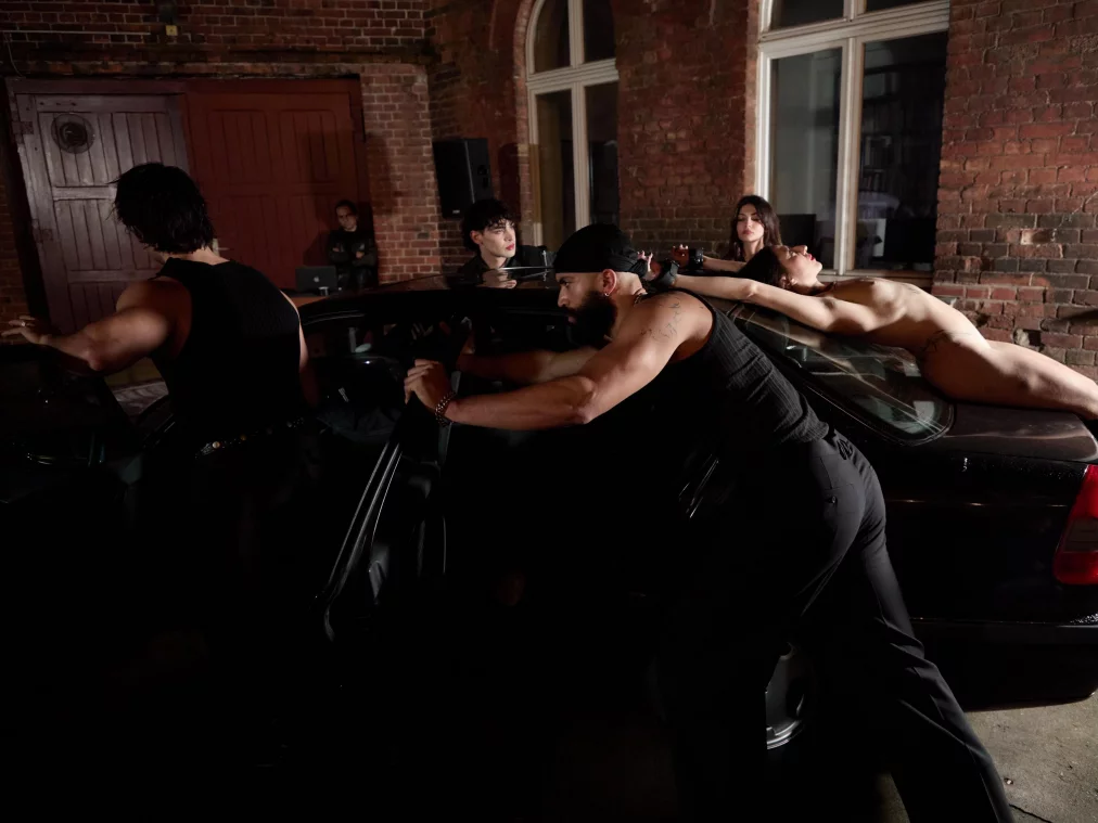 Mehrere Personen lehnen oder liegen auf einem parkenden Auto in einem Innenhof mit Backsteinfassade; weitere Personen befinden sich im Hintergrund.