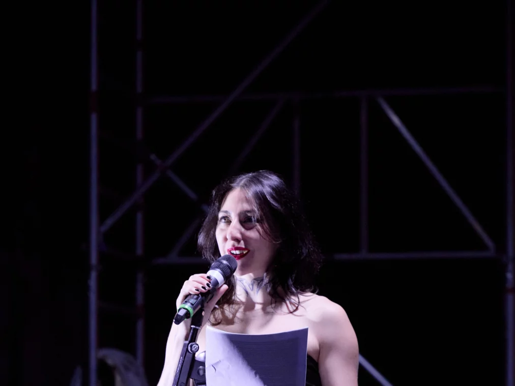 Eine Person im schwarzen Kleid steht auf einer Bühne an einem Mikrofon und liest von einem Blatt Papier; hinter ihr sind Gerüststrukturen und Bühnenlicht sichtbar.