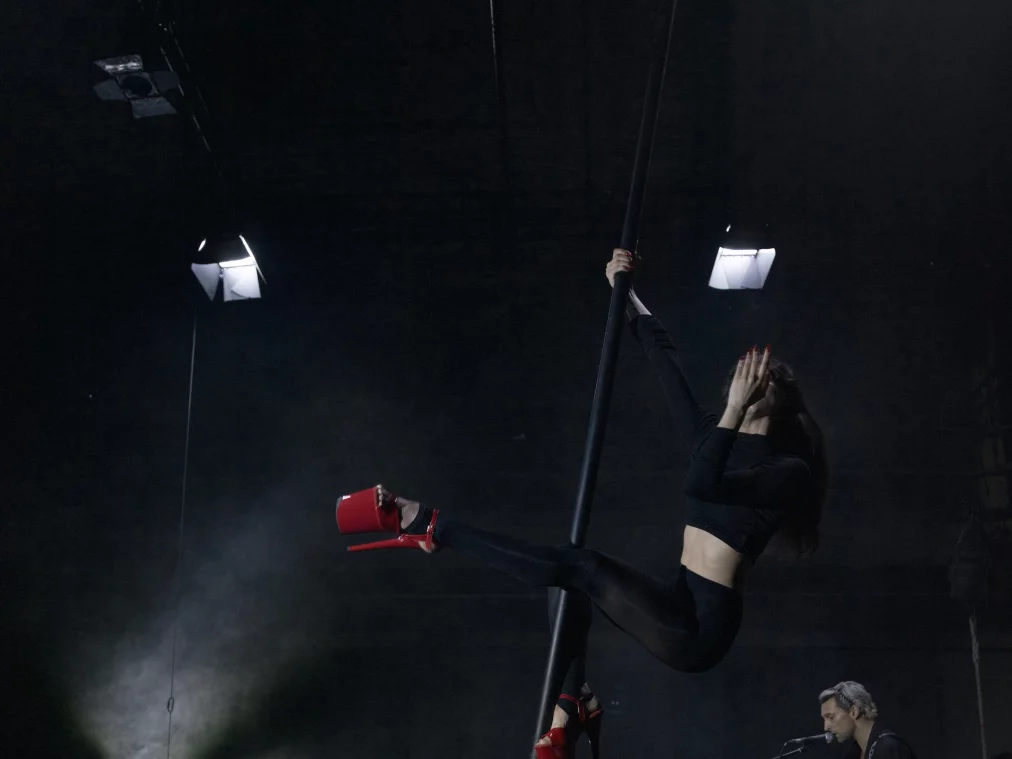 Eine Person hängt an einer vertikalen Stange in der Luft und trägt schwarze Kleidung sowie rote Plateauschuhe; daneben sitzt eine weitere Person auf einem Hocker und spricht oder singt in ein Mikrofon.