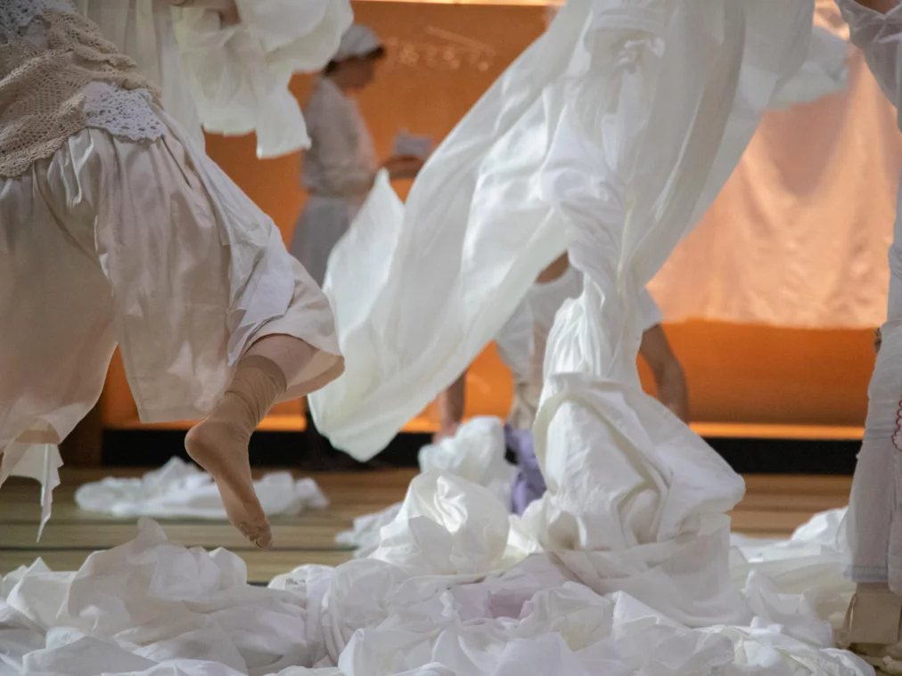 Mehrere Personen in weißen, historischen Kostümen bewegen sich über eine Bühne, während große Mengen weißer Stoffe durch die Luft und über den Boden geworfen sind. Warme orangefarbene Beleuchtung im Hintergrund.