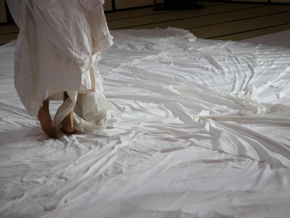 Nahaufnahme von Füßen und Stoffen: Eine Person schreitet barfuß über eine große, ausgebreitete Fläche aus weißen Textilien, die den Boden vollständig bedecken.