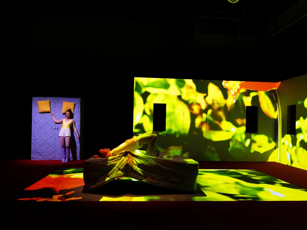 Two performers are on a stage with a red floor. One person lies arched backwards over a bed or mattress, while another stands next to an upright mattress holding a yellow pillow. The walls of the room installation are covered with a bright green, abstract video projection.