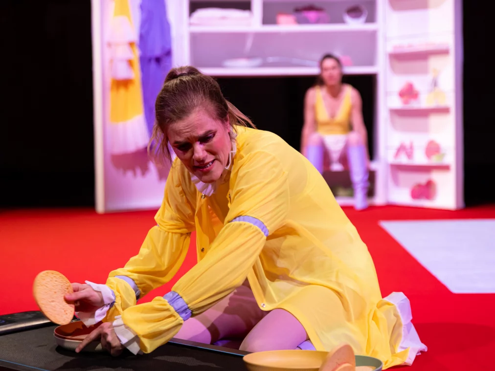 Two performers sit inside an opened, cabinet-like stage prop with shelves. Both wear yellow tops, light shorts, and lilac boots. They sit facing each other in the middle compartment; one person holds a large metal pan and a kitchen utensil. The shelves contain folded fabrics, clothing, food props, and household objects. A yellow garment and a lilac robe hang on the inside of the cabinet doors.