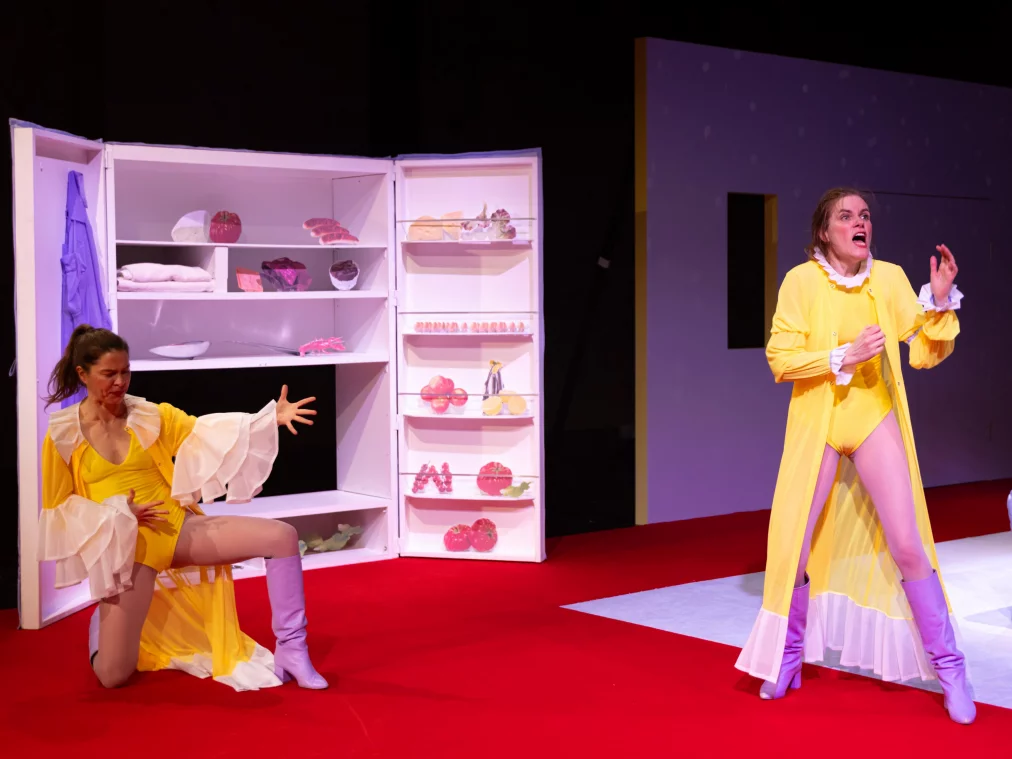 Two performers are on a stage with a red floor next to an opened cabinet-like stage prop. Both wear yellow ruffled costumes and lilac boots. One performer kneels in front of the cabinet, gesturing with outstretched arms, while another performer stands further forward, raising their hands while speaking or singing. A white mattress lies on the stage to the right.