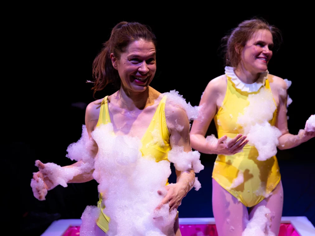 Two persons stand next to each other on a stage in front of a dark background. Both wear yellow sleeveless bodysuits and are partially covered with white foam spread across their upper bodies, arms, and hands. Both persons smile and hold foam in their hands. A pink basin edge is visible behind them at the lower edge of the image.