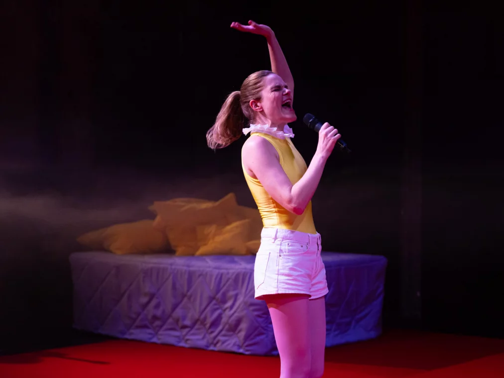Ein*e Performer*in steht auf einer Bühne mit rotem Boden vor einem Bett mit grauer Tagesdecke und gelben Kissen. Die Person trägt ein gelbes ärmelloses Oberteil, eine helle kurze Hose und hält ein Mikrofon. Mit erhobenem Arm singt oder ruft die Person in das Mikrofon, während warmes Bühnenlicht die Szene beleuchtet.