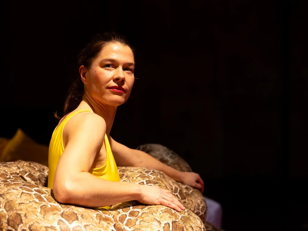 Ein*e Performer*in liegt halb aufgerichtet auf einem Bett oder einer gepolsterten Fläche und lehnt sich an ein großes, schlangenartig gemustertes Kissen oder Objekt. Die Person trägt ein gelbes ärmelloses Oberteil, blickt ruhig zur Seite und wird von warmem Bühnenlicht ausgeleuchtet.