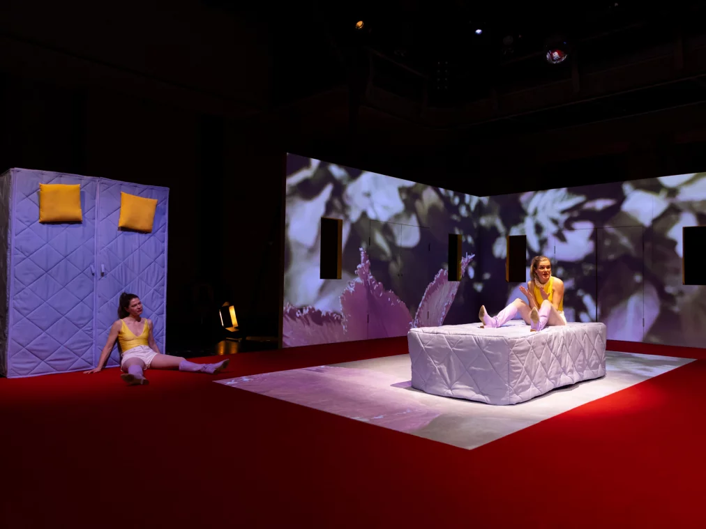 Zwei Performer*innen befinden sich auf einer Bühne mit rotem Boden vor einer weißen Rauminstallation mit floralen Videoprojektionen. Eine Person sitzt auf einer weißen Matratze in der Bühnenmitte, trägt ein gelbes ärmelloses Oberteil, eine helle kurze Hose und lilafarbene Stiefel und gestikuliert mit den Händen. Eine zweite Person sitzt links am Boden, lehnt an einer aufrecht stehenden Matratze mit zwei gelben Kissen.