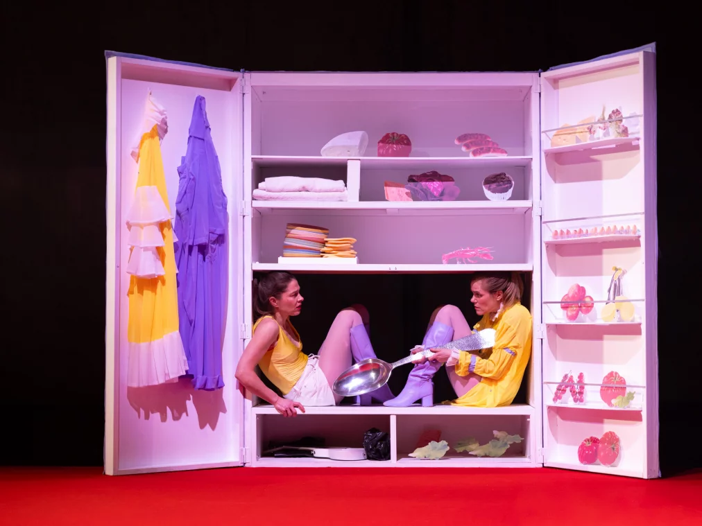 Zwei Performer*innen sitzen in einem geöffneten, schrankartigen Bühnenobjekt mit Regalfächern. Beide tragen gelbe Oberteile, helle kurze Hosen und lilafarbene Stiefel. Sie sitzen einander gegenüber im mittleren Fach; eine Person hält eine große Metallpfanne und einen Küchenutensil. In den Regalen befinden sich gefaltete Stoffe, Kleidung, Lebensmittelattrappen und Haushaltsgegenstände. An der Innenseite der Türen hängen ein gelbes Kleidungsstück und ein lilafarbenes Gewand.