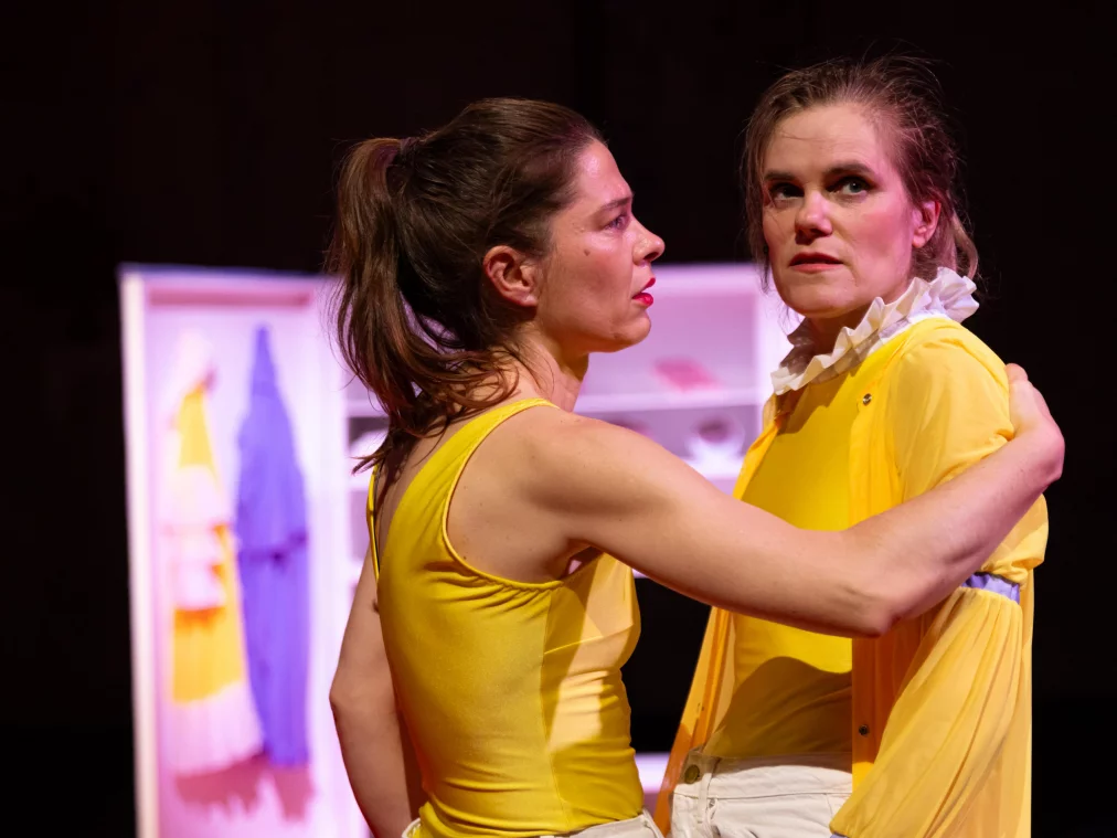 Zwei Performer*innen stehen dicht beieinander und halten sich um die Schultern. Beide tragen gelbe Oberteile und helle kurze Hosen; ein*e Performer*in trägt zusätzlich ein transparentes gelbes Überkleid mit Rüschenkragen. Beide blicken in unterschiedliche Richtungen, während sie eng miteinander verbunden bleiben.