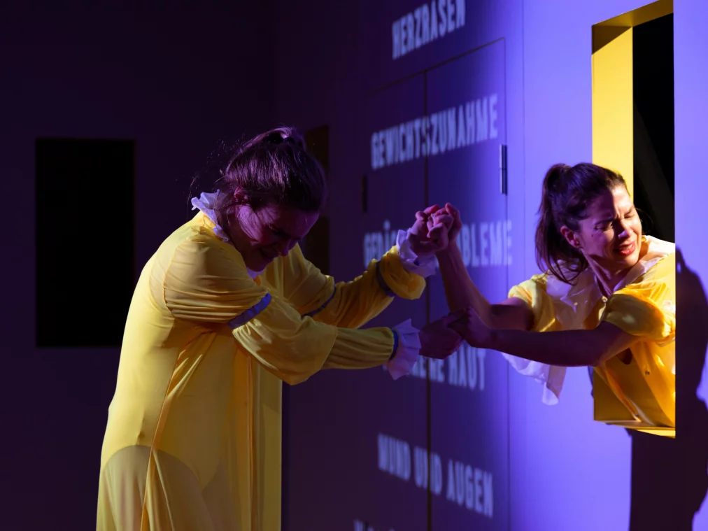 Zwei Performer*innen stehen an einer Wand der Rauminstallation neben einer rechteckigen Öffnung. Sie halten sich an den Händen und drücken gegeneinander, während ein*e Performer*in durch die Öffnung nach außen ragt. Auf der Wand erscheinen projizierte Textzeilen. Beide tragen gelbe Kostüme mit Rüschen.