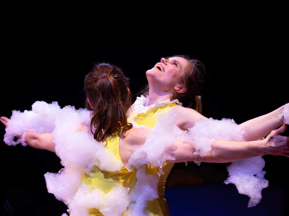 Zwei Performer*innen stehen Rücken an Rücken mit ausgestreckten Armen. Beide tragen gelbe Oberteile und sind teilweise von weißem Schaum bedeckt, der sich über Schultern, Arme und Rücken verteilt. Eine Person neigt den Kopf nach hinten und lächelt, während Bühnenlicht die Szene hervorhebt.