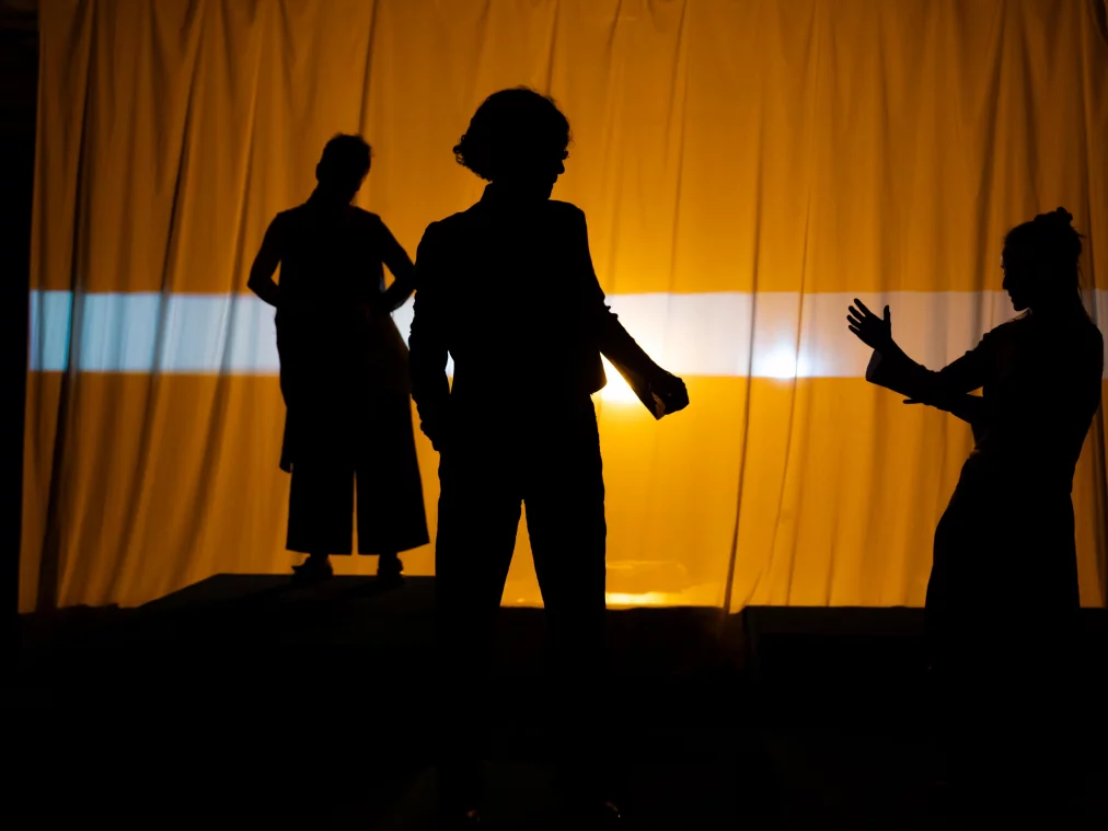 Drei Personen stehen als Silhouetten vor einem gelb beleuchteten Vorhang. Ihre Körper sind im Gegenlicht nur als dunkle Umrisse erkennbar; eine Person hebt eine Hand zu einer Geste.©MayraWallraff-45