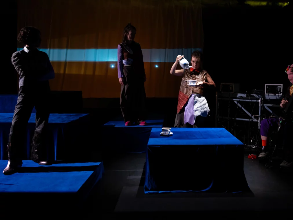 Mehrere Personen verteilen sich auf Podesten und um einen Tisch. Eine Person gießt Tee aus einer weißen Kanne in eine Tasse. Rechts sitzt eine weitere Person an einem Instrument oder technischen Gerät. Das Bühnenlicht ist dunkel mit blau-gelben Kontrasten.
